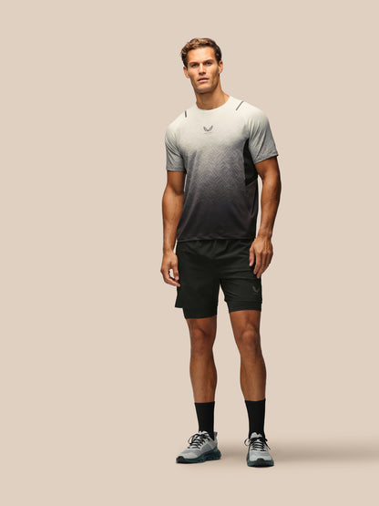 A male model stands against a solid beige studio background wearing a short-sleeve athletic t-shirt with a gray-to-black gradient, black running shorts, black socks, and gray running shoes.