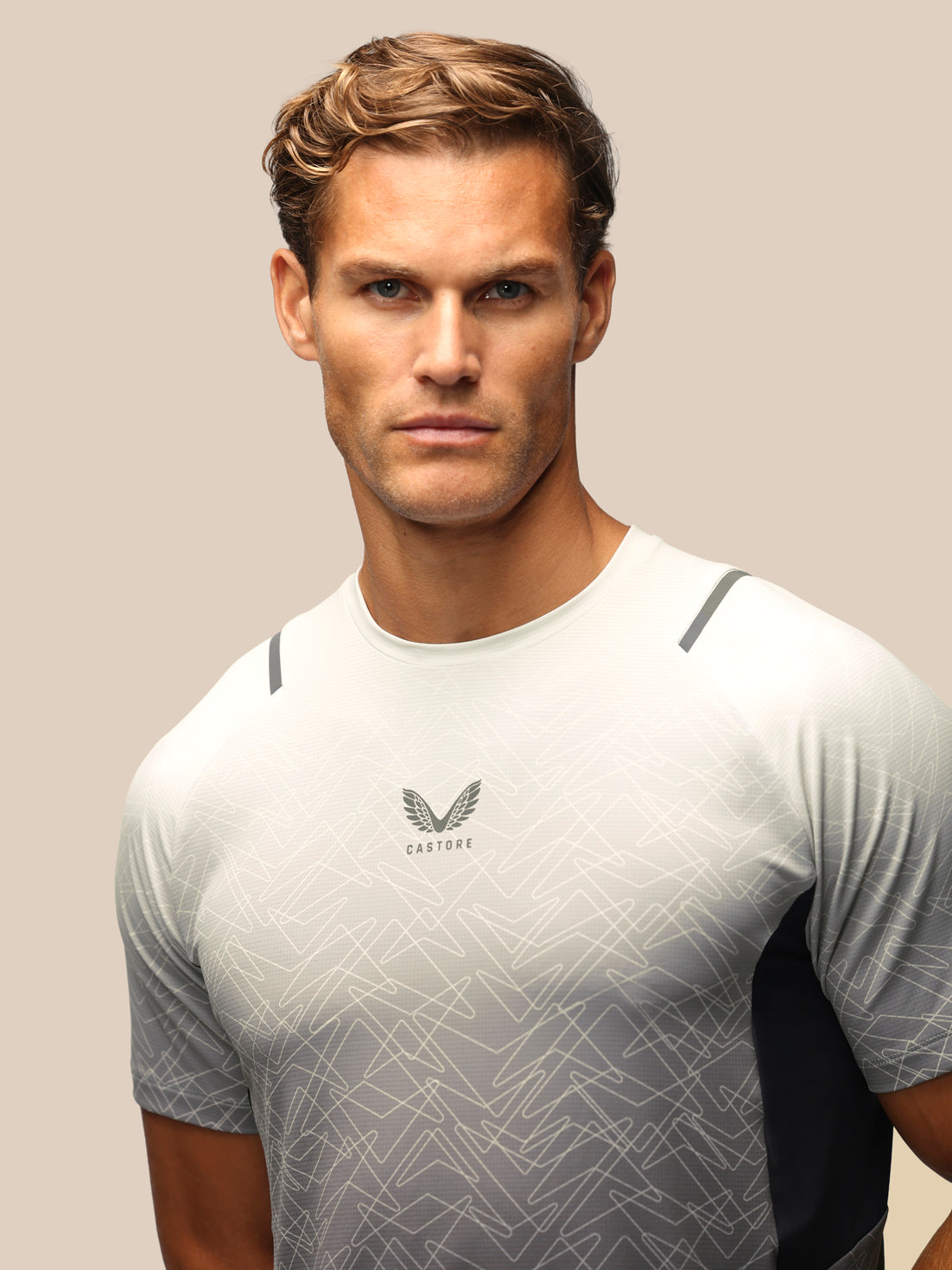 A man wearing a light gray and white athletic t-shirt with a subtle geometric pattern and a winged logo on the chest that reads 'CASTORE'. The background is a solid, warm beige studio backdrop.