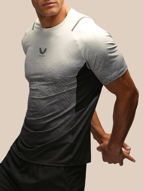 A close-up of a man wearing a short-sleeve athletic t-shirt with a gray and black gradient design, featuring a subtle geometric pattern and a black wing logo on the chest.