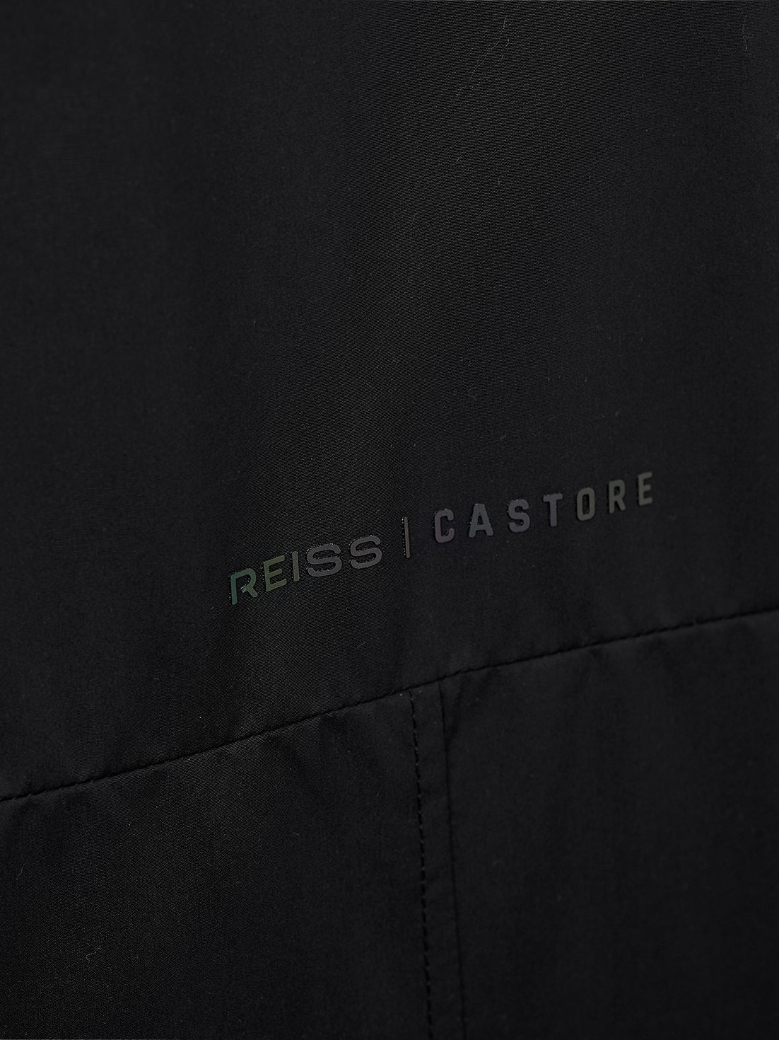 Close-up of a black garment with the REISS | CASTORE logo printed across the chest in a subtle iridescent tone.