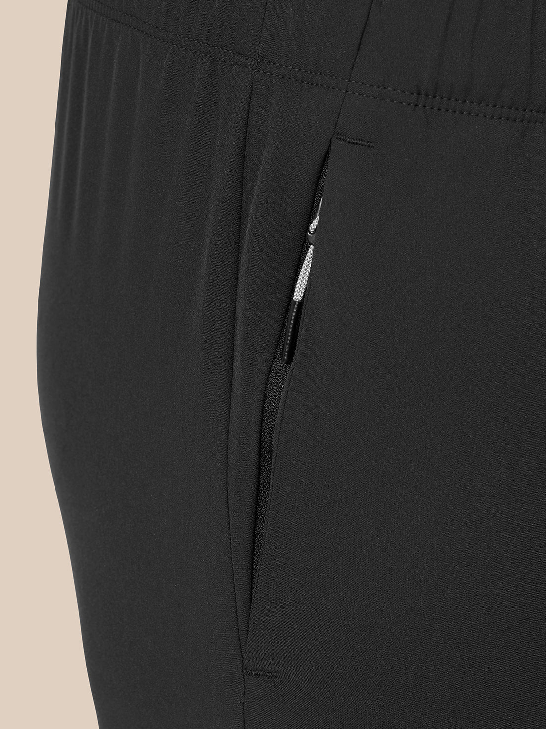 Close-up of black athletic pants showing a side zippered pocket with a black zipper pull and a small white accent, photographed against a light beige background.