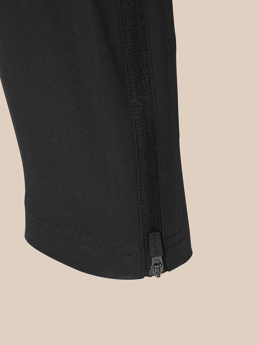 Close-up of a black garment leg with a vertical zippered detail near the hem, shown against a solid beige background.