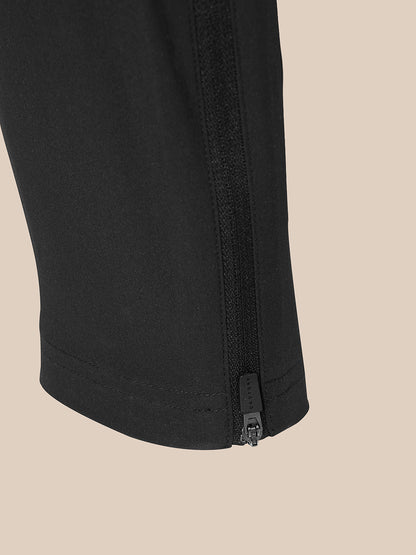 Close-up of a black garment leg with a vertical zippered detail near the hem, shown against a solid beige background.