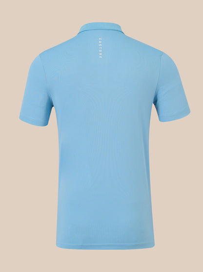 A light blue short-sleeve polo shirt shown from the back, featuring a vertical 'CASTORE' logo printed in white near the collar, set against a solid beige background.