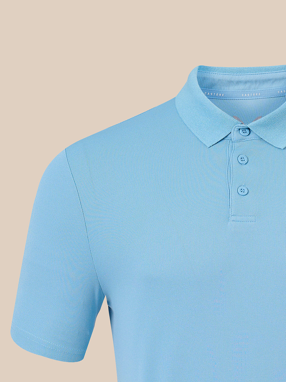 A close-up of a light blue short-sleeve polo shirt on a mannequin or torso form, showing the collar, three-button placket, and ribbed fabric against a solid beige background.