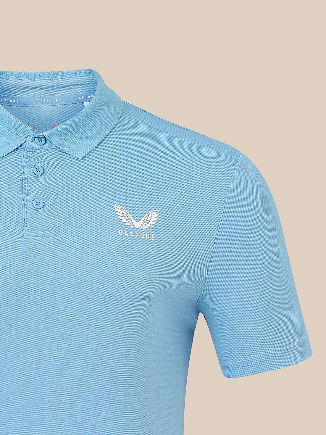 A light blue short-sleeve polo shirt with a white embroidered winged logo and the word 'CASTORE' on the left chest, shown against a solid beige background.
