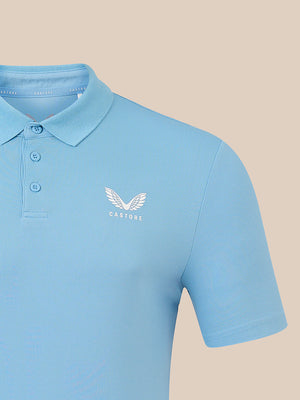 A light blue short-sleeve polo shirt with a white embroidered winged logo and the word 'CASTORE' on the left chest, shown against a solid beige background.
