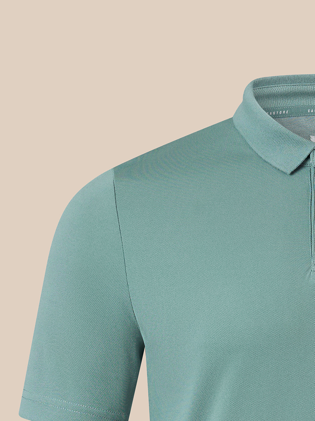 Close-up of a short-sleeve polo shirt in a muted teal-green color, showing the collar, shoulder seam, and sleeve on a plain beige background.