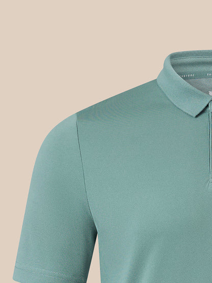 Close-up of a short-sleeve polo shirt in a muted teal-green color, showing the collar, shoulder seam, and sleeve on a plain beige background.