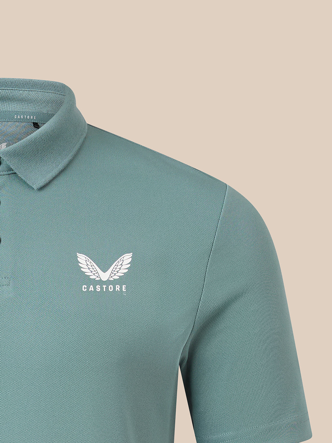 Close-up of a short-sleeve polo shirt in a muted teal-green color, featuring a white embroidered winged logo and the brand name 'CASTORE' on the left chest, set against a solid beige background.