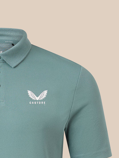Close-up of a short-sleeve polo shirt in a muted teal-green color, featuring a white embroidered winged logo and the brand name 'CASTORE' on the left chest, set against a solid beige background.