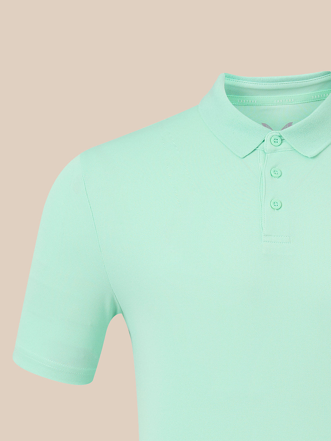 A close-up of a mint green short-sleeve polo shirt on a solid pale peach-beige background.