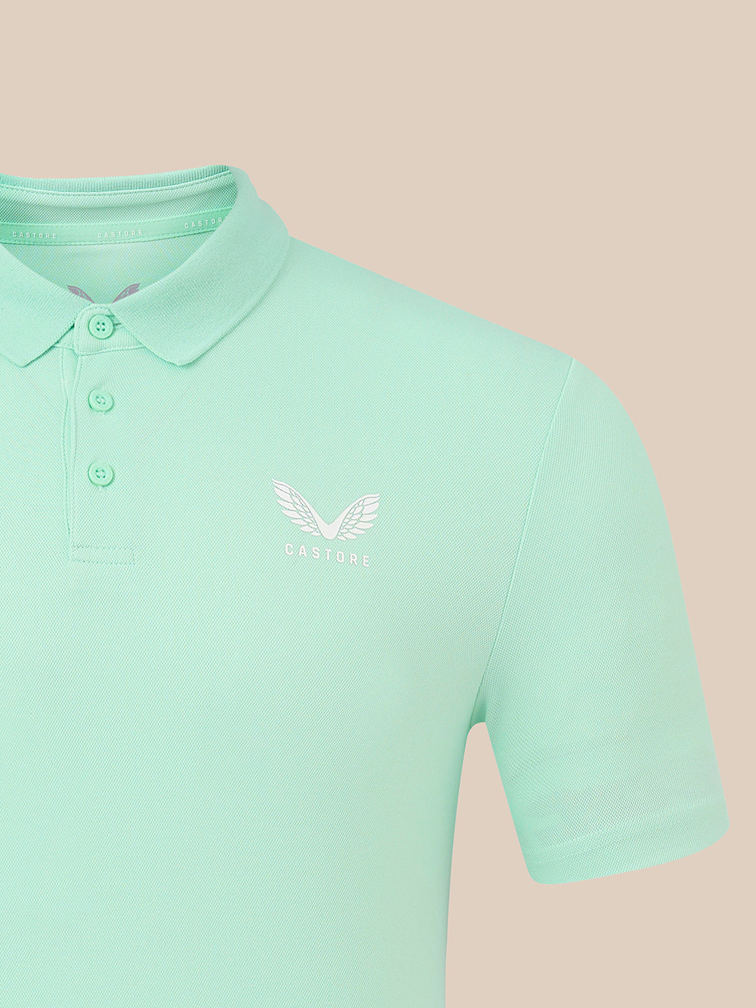 A mint green short-sleeve polo shirt with a white embroidered winged logo and the word 'CASTORE' on the left chest, shown against a solid pale peach background.