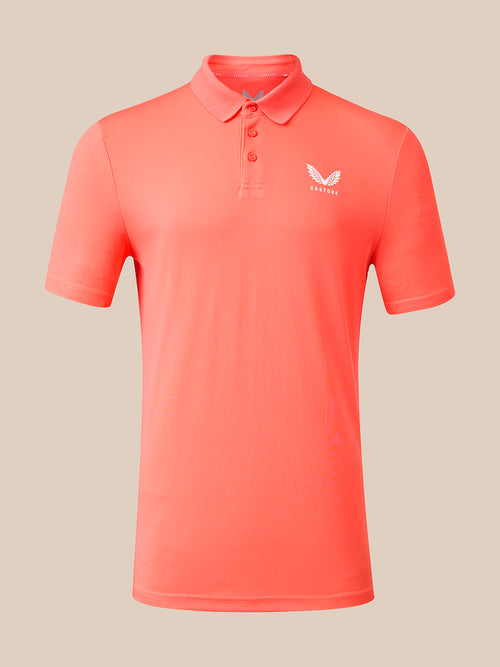 A coral pink short-sleeve polo shirt with a three-button placket and a small white wing logo on the left chest, displayed on a neutral beige background.