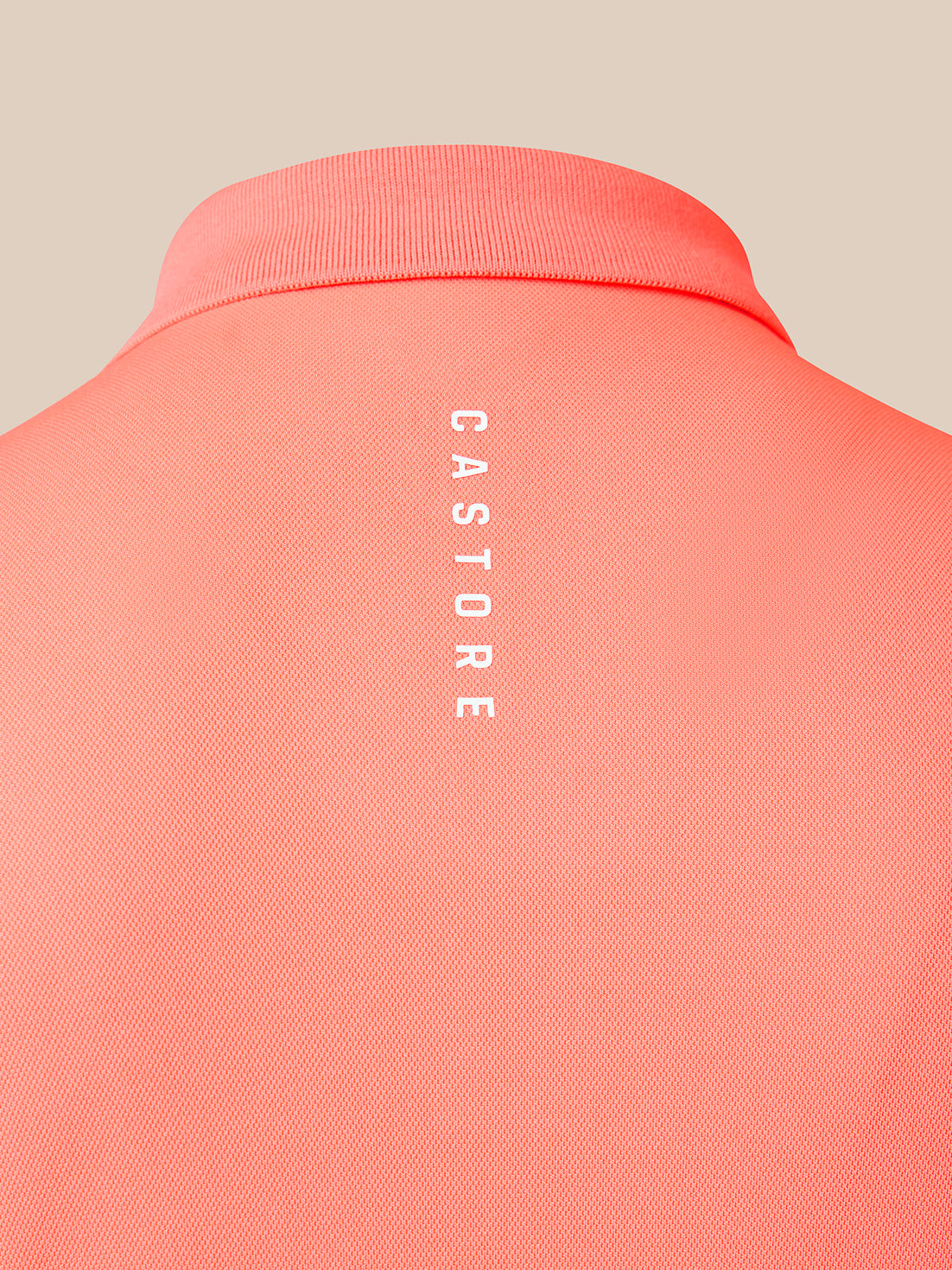 Close-up of the back of a coral pink polo shirt with vertical white 'CASTORE' branding printed on the center back, set against a solid beige background.
