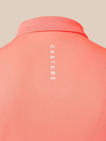 Close-up of the back of a coral pink polo shirt with vertical white 'CASTORE' branding printed on the center back, set against a solid beige background.