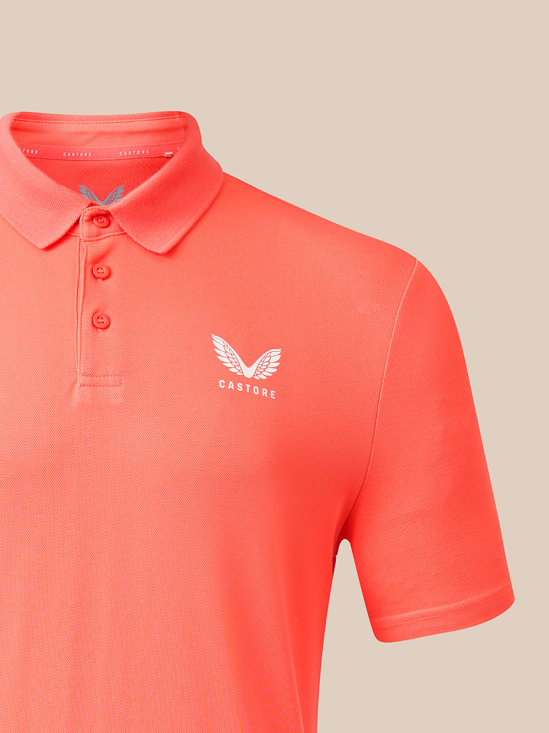 A close-up of a coral orange short-sleeve polo shirt with a three-button placket and a white embroidered winged logo with the word 'CASTORE' on the left chest, set against a solid beige background.