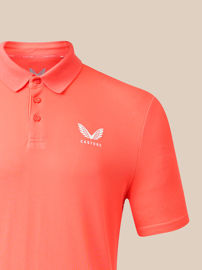 A close-up of a coral orange short-sleeve polo shirt with a three-button placket and a white embroidered winged logo with the word 'CASTORE' on the left chest, set against a solid beige background.