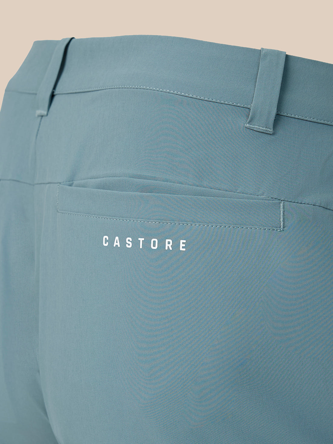 Close-up of the back waistband and pocket area of dusty blue trousers, featuring a back pocket with the brand name 'CASTORE' printed in white.