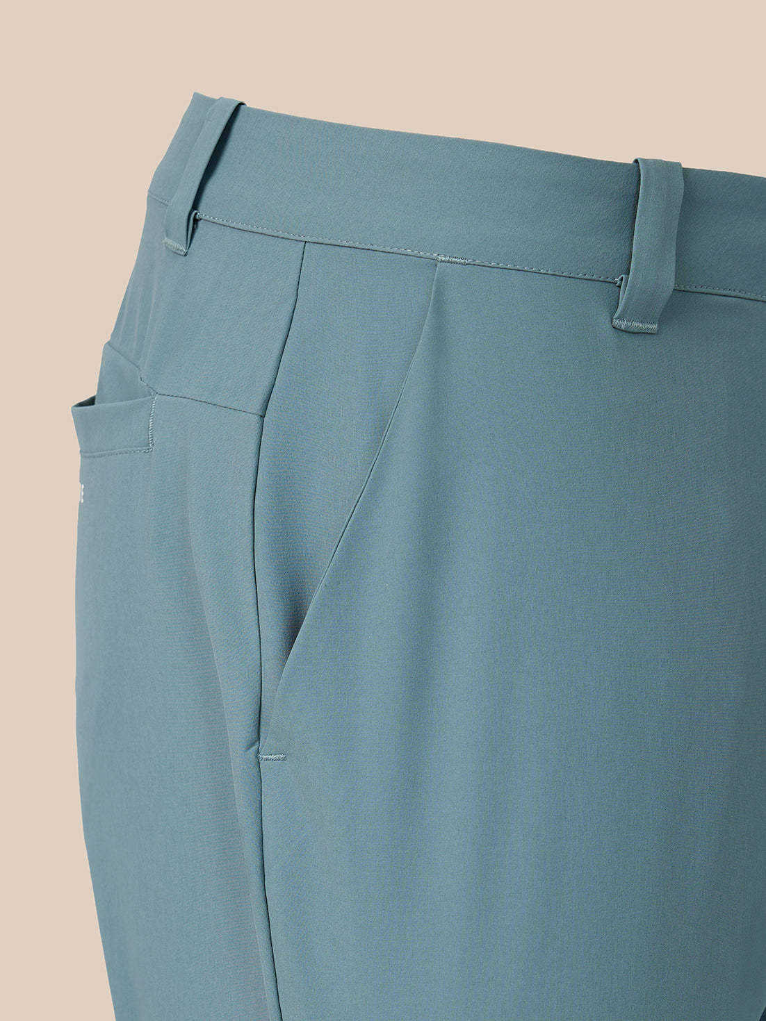 Close-up of a pair of tailored trousers in a muted dusty blue color, showing the waistband, belt loops, side pocket, and front crease against a solid beige background.