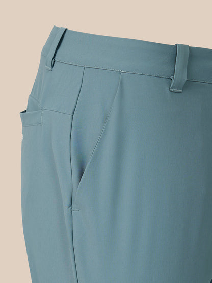 Close-up of a pair of tailored trousers in a muted dusty blue color, showing the waistband, belt loops, side pocket, and front crease against a solid beige background.