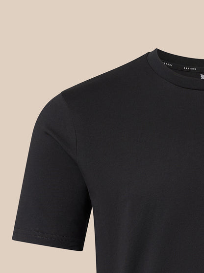 Close-up of a black short-sleeve crewneck t-shirt on a light beige background, showing the inner collar label with the brand name CASTORE
