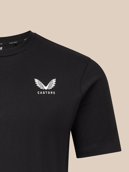 Close-up of a black short-sleeve crewneck t-shirt featuring a white embroidered winged logo and the brand name 'CASTORE' on the left chest, set against a solid beige background.