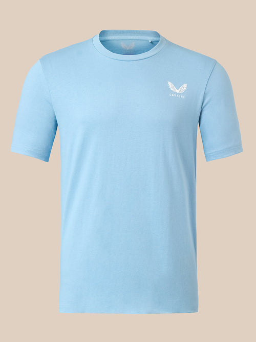 A light blue short-sleeve crewneck t-shirt with a small white winged logo on the left chest, displayed on a solid beige background.