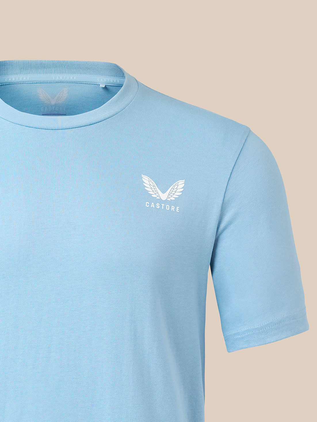 A light blue short-sleeve crewneck t-shirt with a white winged logo and the word 'CASTORE' printed on the left chest, shown against a solid beige background.