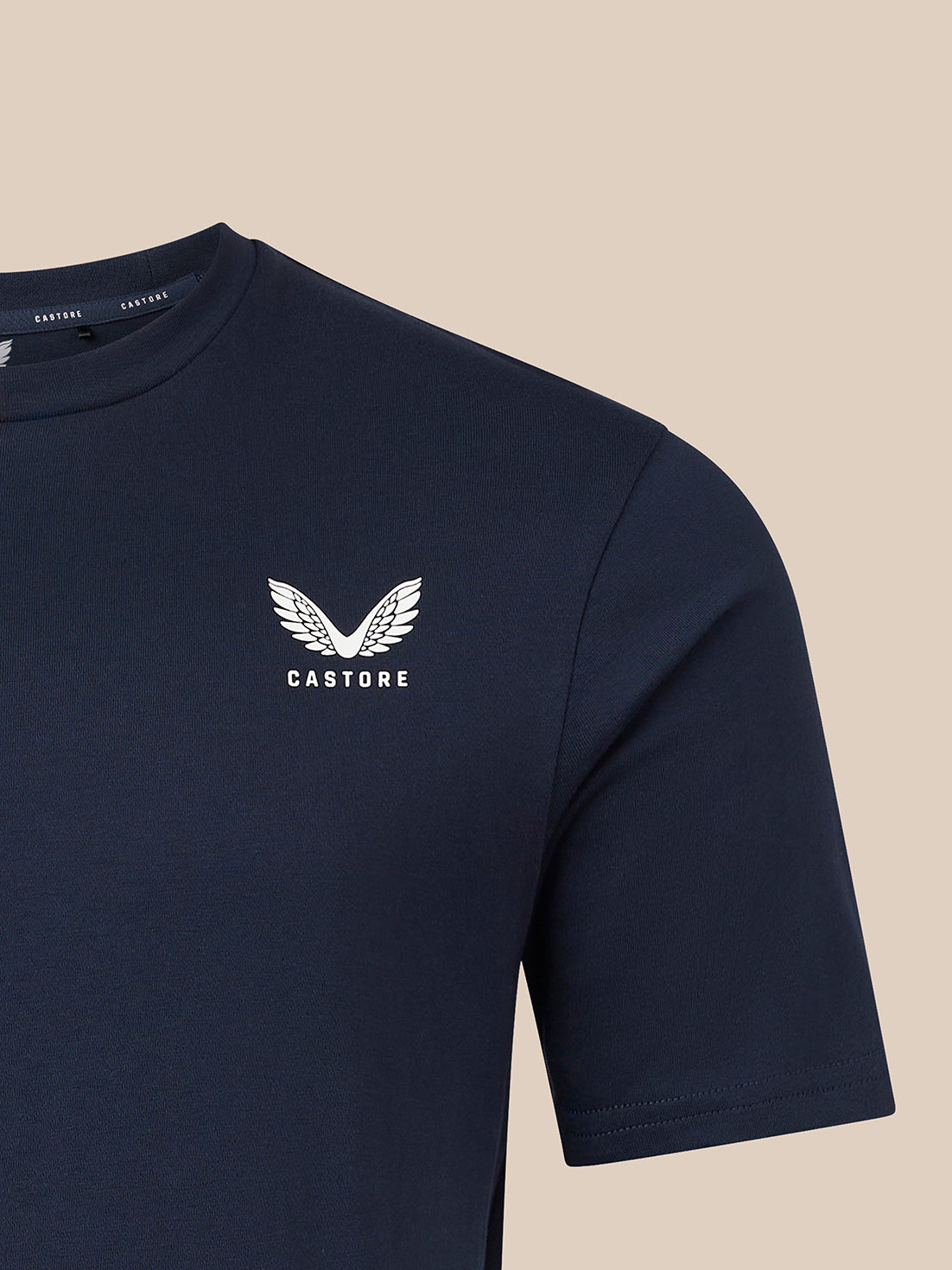 A close-up of a navy blue short-sleeve t-shirt with a white Castore wing logo and brand name printed on the left chest, set against a solid beige background.