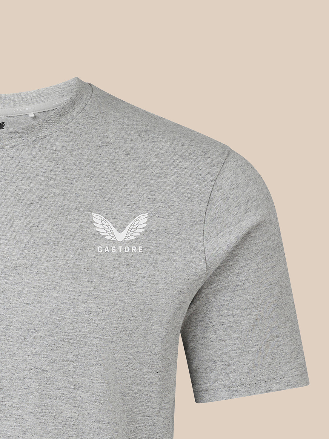 A close-up of a heather gray crewneck t-shirt featuring a white winged logo and the word 'CASTORE' printed on the left chest, set against a solid beige background.