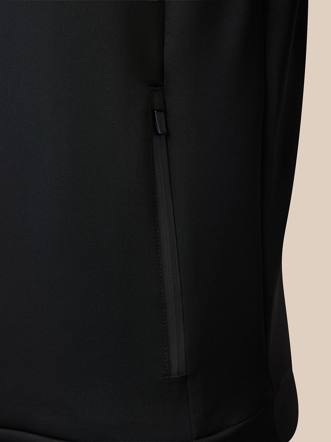 Close-up of a black zippered pocket on a black garment, with a small portion of a light beige background visible on the right side.