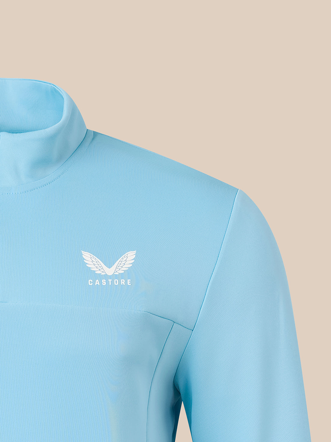 A close-up of a light blue athletic long-sleeve top with a mock neck collar, featuring a white winged logo and the brand name 'CASTORE' on the left chest, set against a solid beige background.