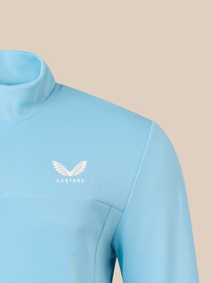 A close-up of a light blue athletic long-sleeve top with a mock neck collar, featuring a white winged logo and the brand name 'CASTORE' on the left chest, set against a solid beige background.