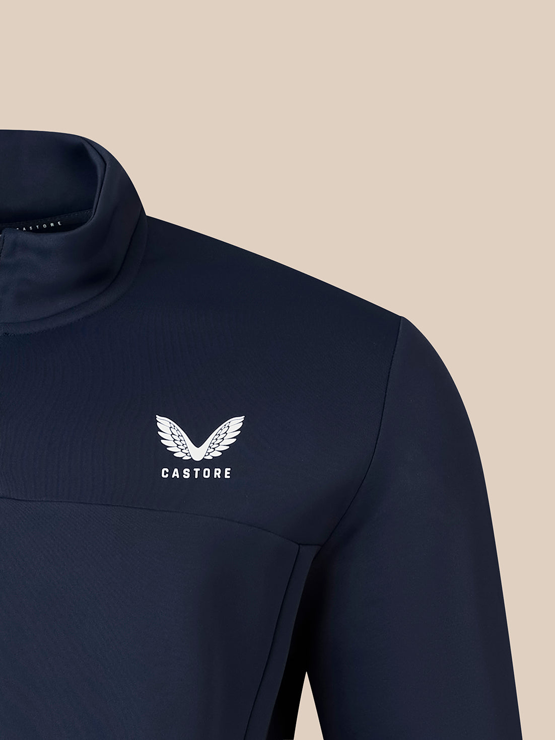 Close-up of a navy athletic quarter-zip top with a white Castore wing logo and text on the chest, set against a solid beige background.