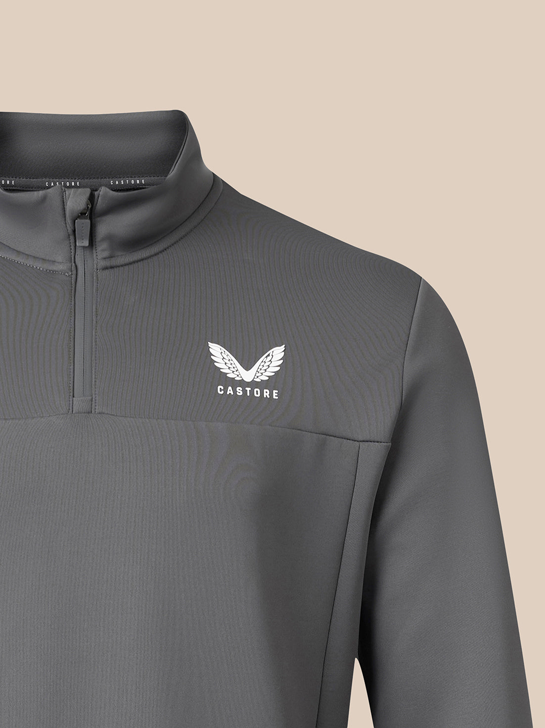 Close-up of a dark gray quarter-zip pullover with a white winged logo and the word 'CASTORE' on the left chest, set against a solid beige background.