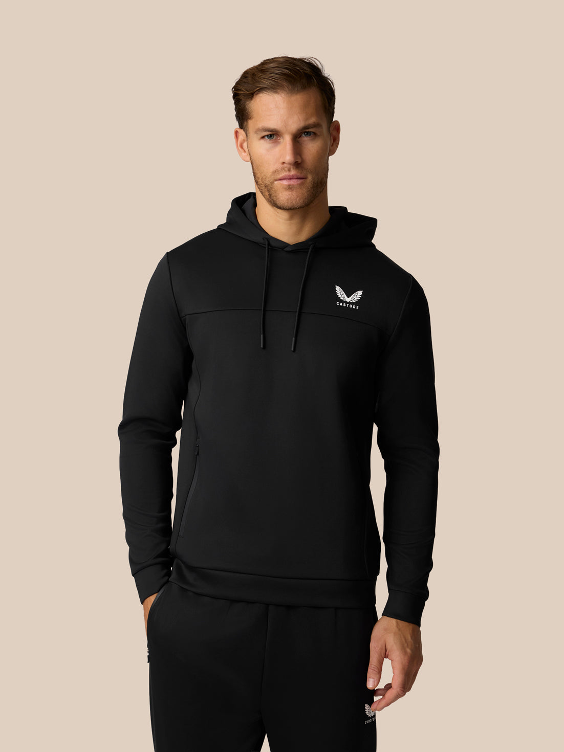 A man wearing a black pullover hoodie with a small white wing logo on the left chest, paired with black sweatpants, set against a solid beige background.