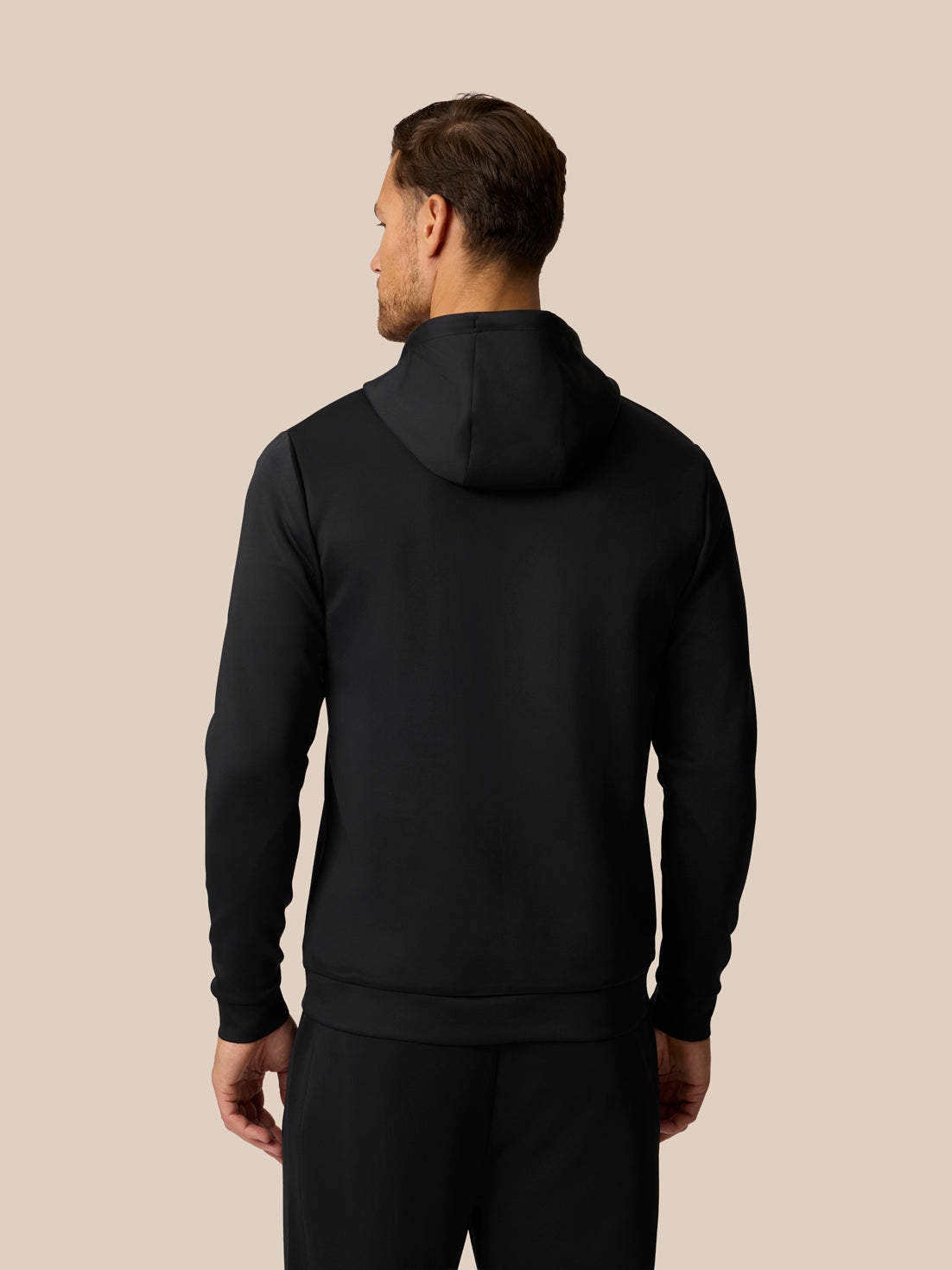 A man viewed from the back wearing a black pullover hoodie and matching black pants against a solid beige studio background.