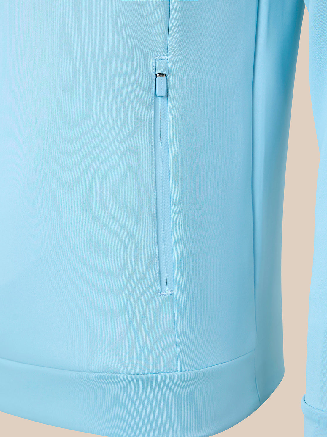 Close-up of a light blue athletic jacket featuring a vertical zippered pocket on the front, set against a solid beige background.