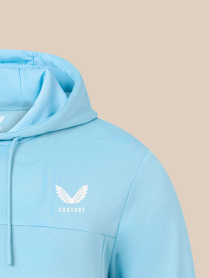 A close-up of a light blue hooded sweatshirt with a white winged logo and the word 'CASTORE' printed on the chest, set against a solid beige background.