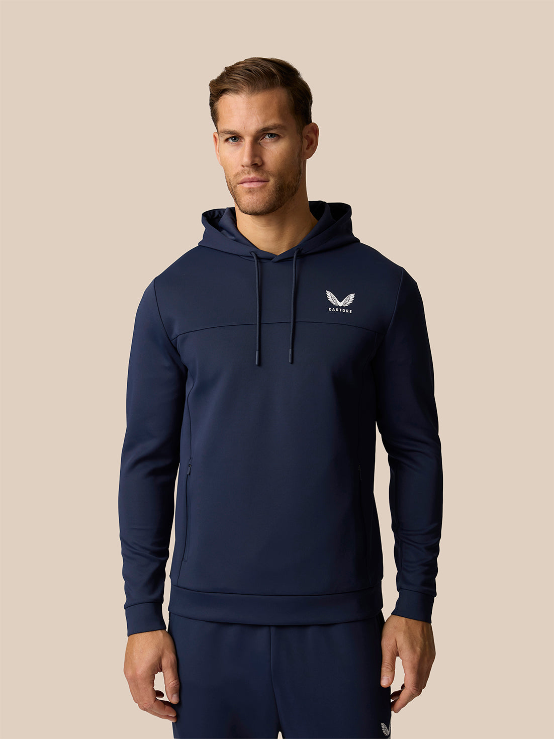 A man wearing a navy blue pullover hoodie with a white winged logo on the left chest, paired with matching navy pants, set against a solid beige background.