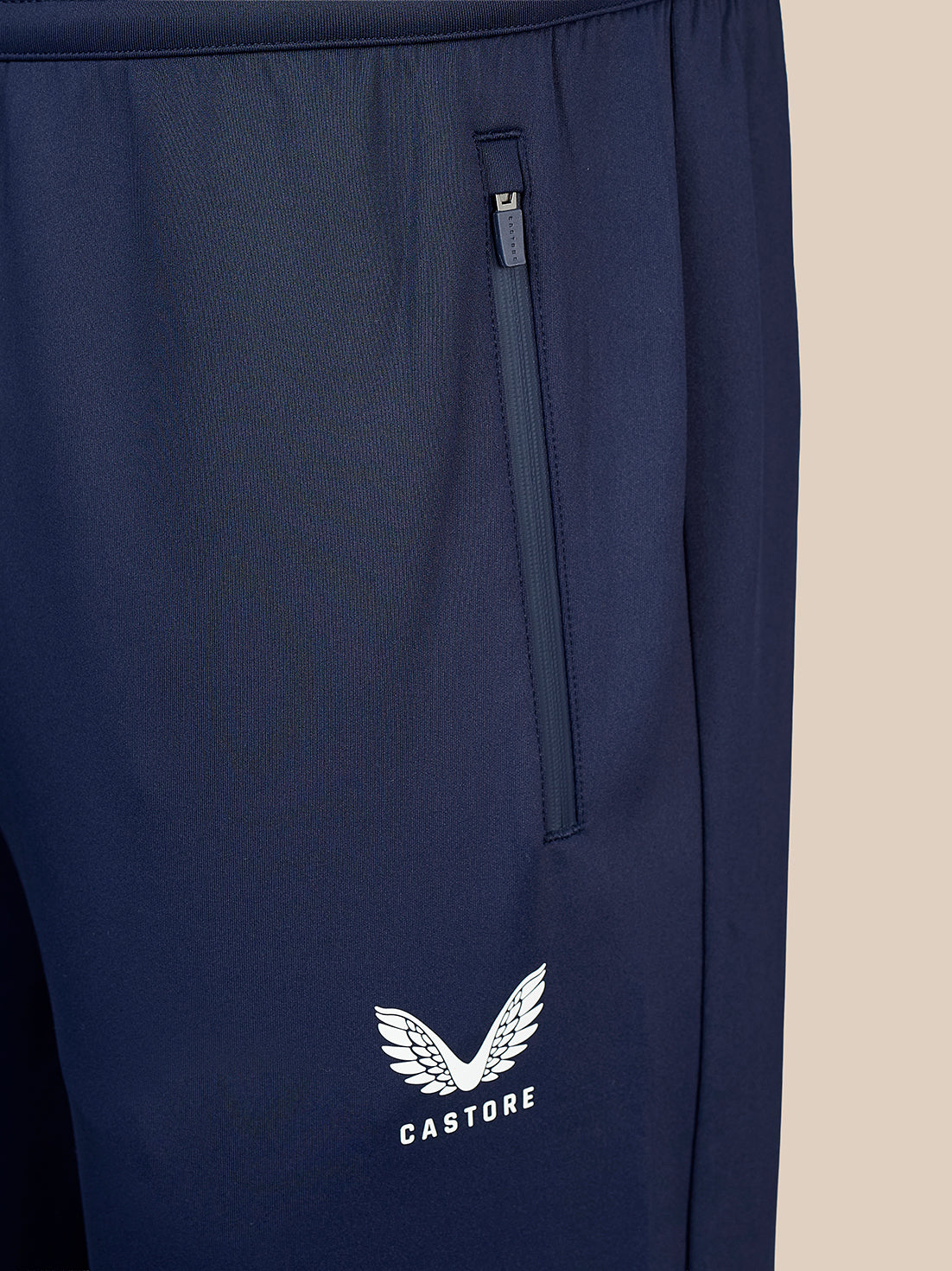 Close-up of navy blue athletic pants featuring a vertical zippered pocket and a white Castore wing logo on the lower leg, set against a solid beige background.
