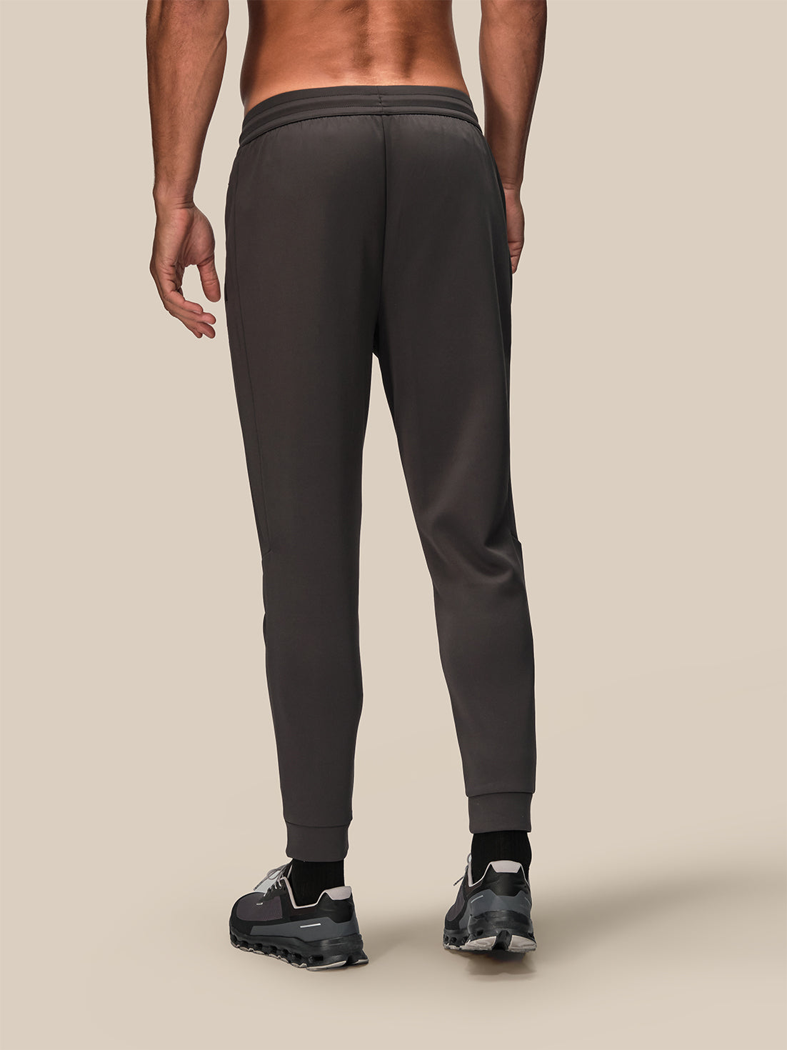 Rear view of a man wearing dark gray jogger sweatpants and black sneakers against a solid beige background.