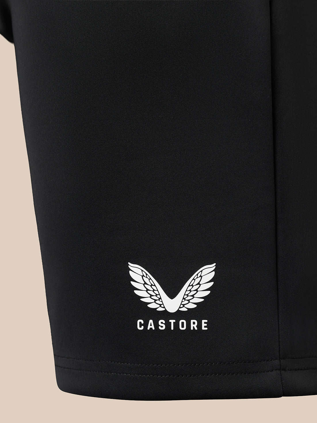 Close-up of a black garment leg featuring a white embroidered wing logo and the brand name 'CASTORE' near the hem, set against a light beige background.