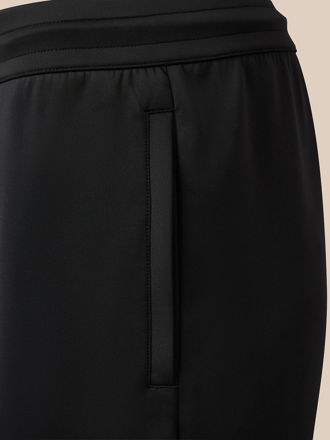 Close-up of black athletic pants showing a side seam, waistband, and a vertical zippered pocket on a light beige background.