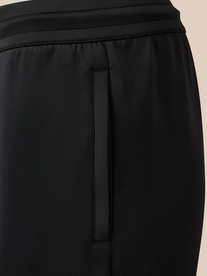 Close-up of black athletic pants showing a side seam, waistband, and a vertical zippered pocket on a light beige background.