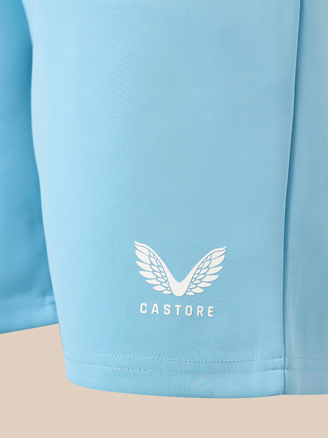 Close-up of a light blue athletic shorts leg featuring a white embroidered wing logo and the brand name 'CASTORE' printed below it.