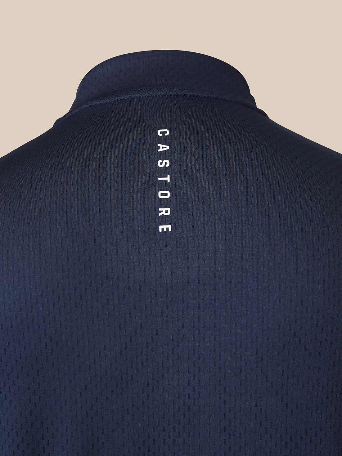 Close-up back view of a dark navy textured polo shirt with the brand name 'CASTORE' printed vertically in white on the center of the back, set against a solid beige background.