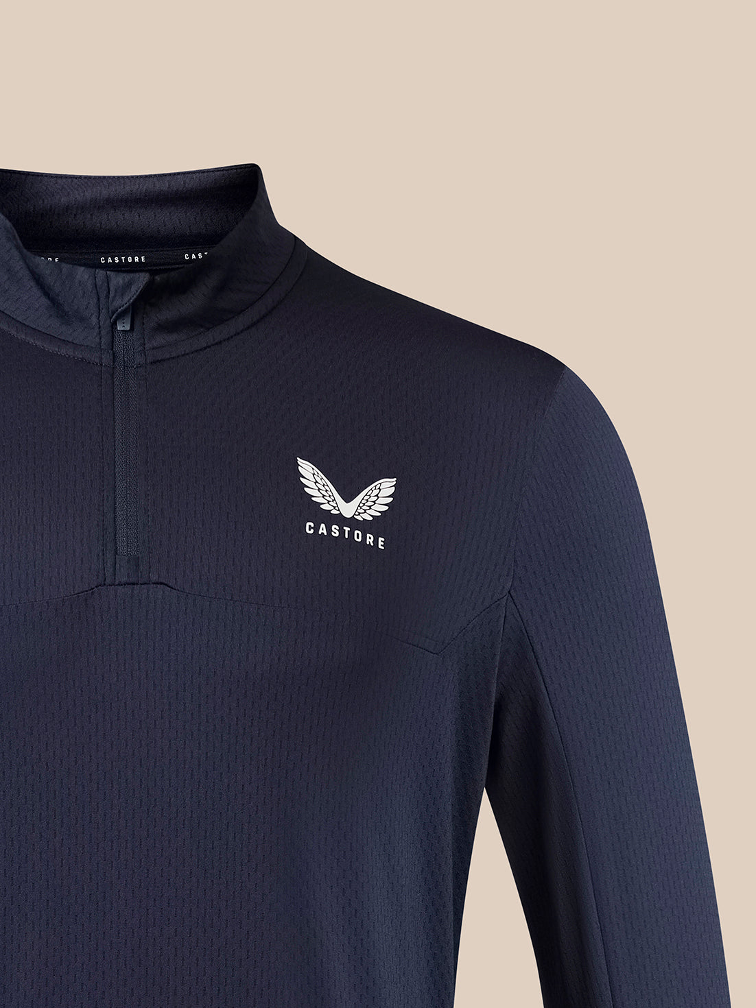 A close-up of a dark navy ribbed long-sleeve quarter-zip athletic top with a white winged logo and the brand name 'CASTORE' on the left chest, set against a solid beige background.