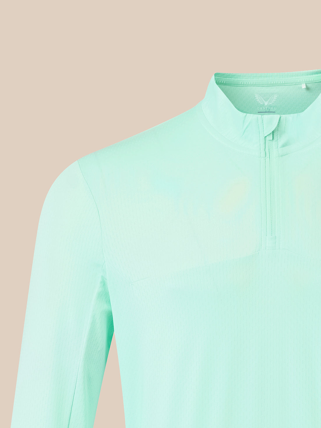 A close-up of a mint green long-sleeve athletic top with a mock neck and half-zip, shown against a solid pale peach background.